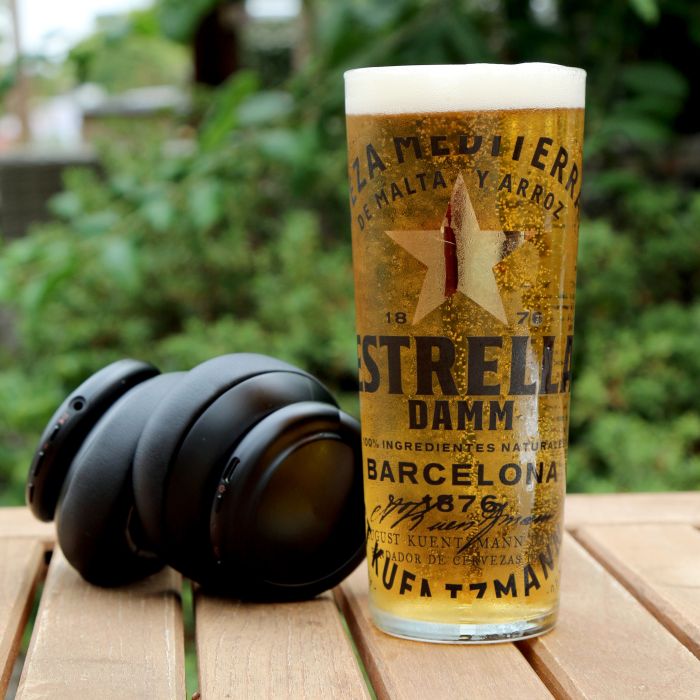 An Estrella Damm pint on a pub garden table and headphones next to the pint. The background is greenery; blurred.